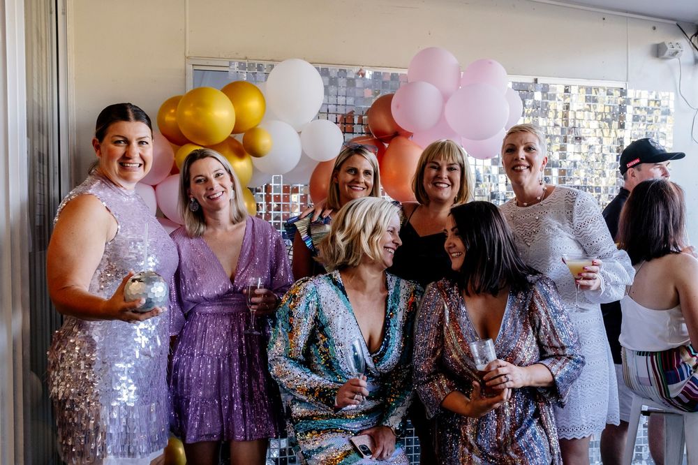 A group of party guests in formal attire gather at an event with colorful balloons and decorations.