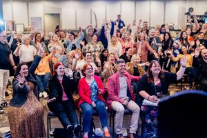 A large enthusiastic crowd of people sitting in rows at an indoor event with raised hands and smiling expressions.