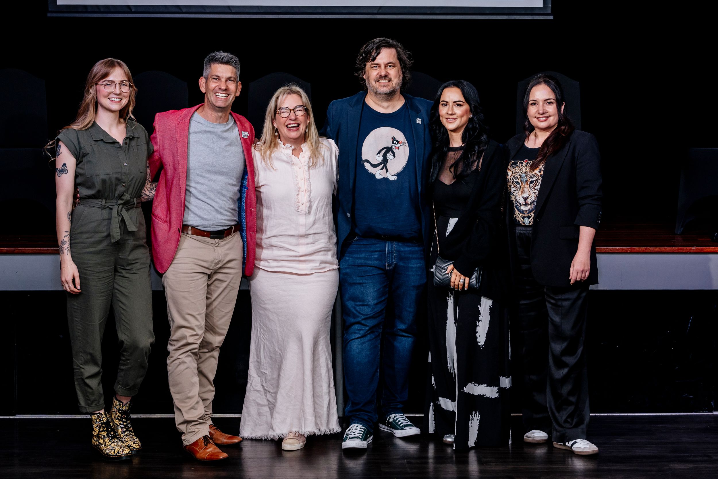 A group of people stand together in casual attire on a dark stage or performance space.