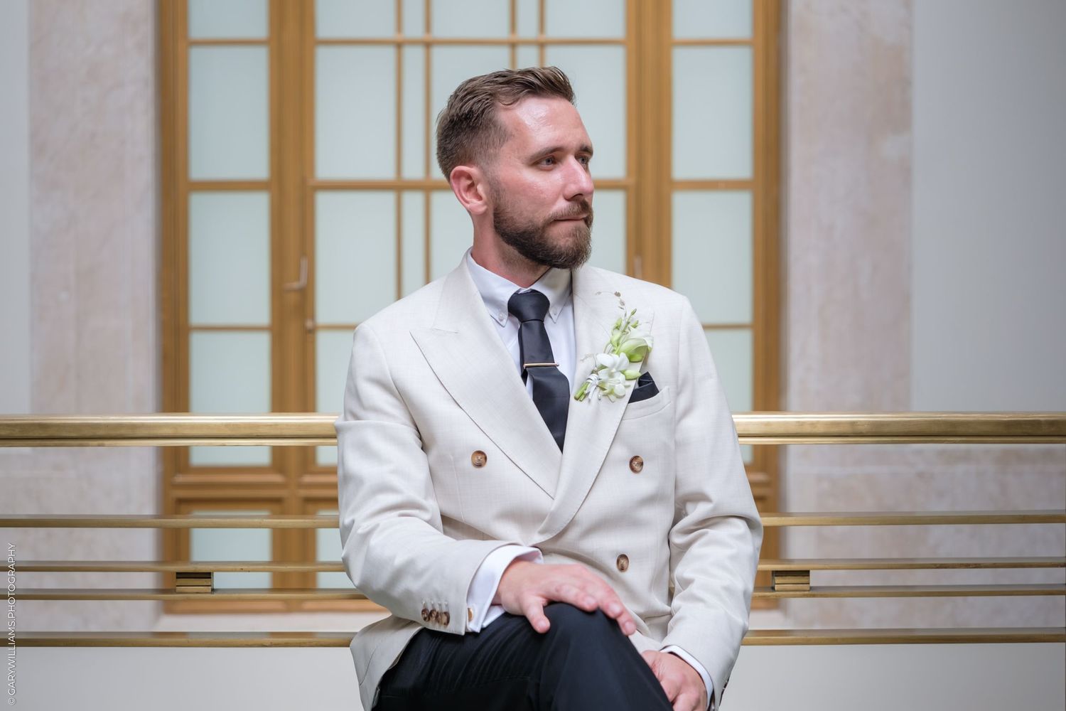 A person in a white tuxedo jacket looks off to the side while standing against an elegant architectural backdrop.