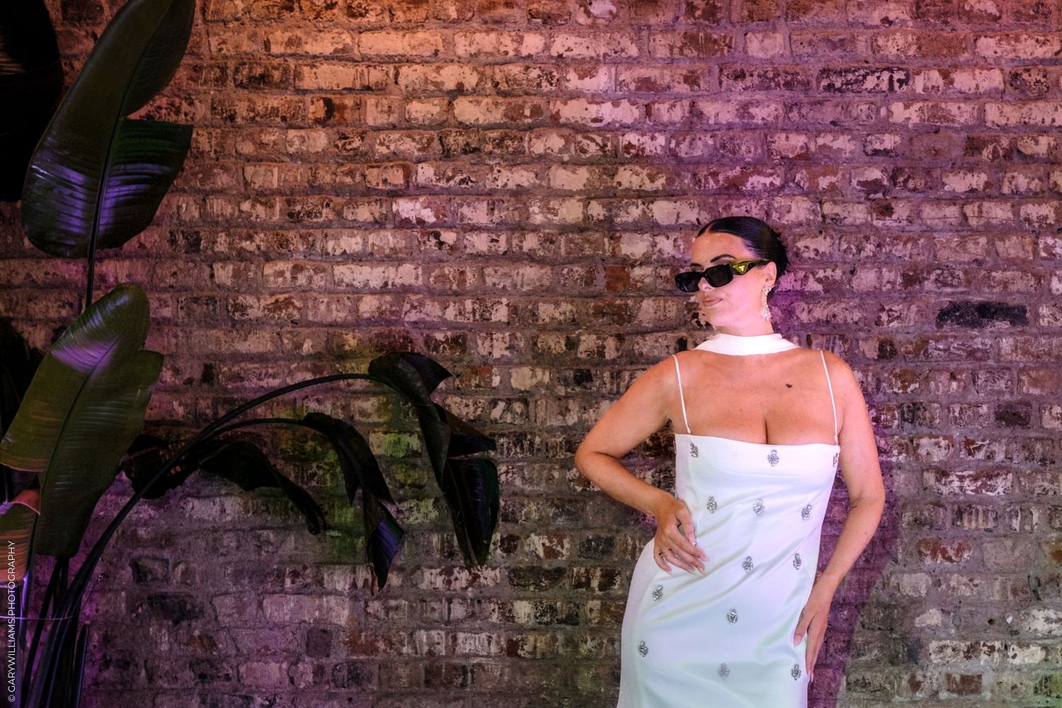 Seven wedding portraits are displayed against a rustic brick wall with green foliage accents.