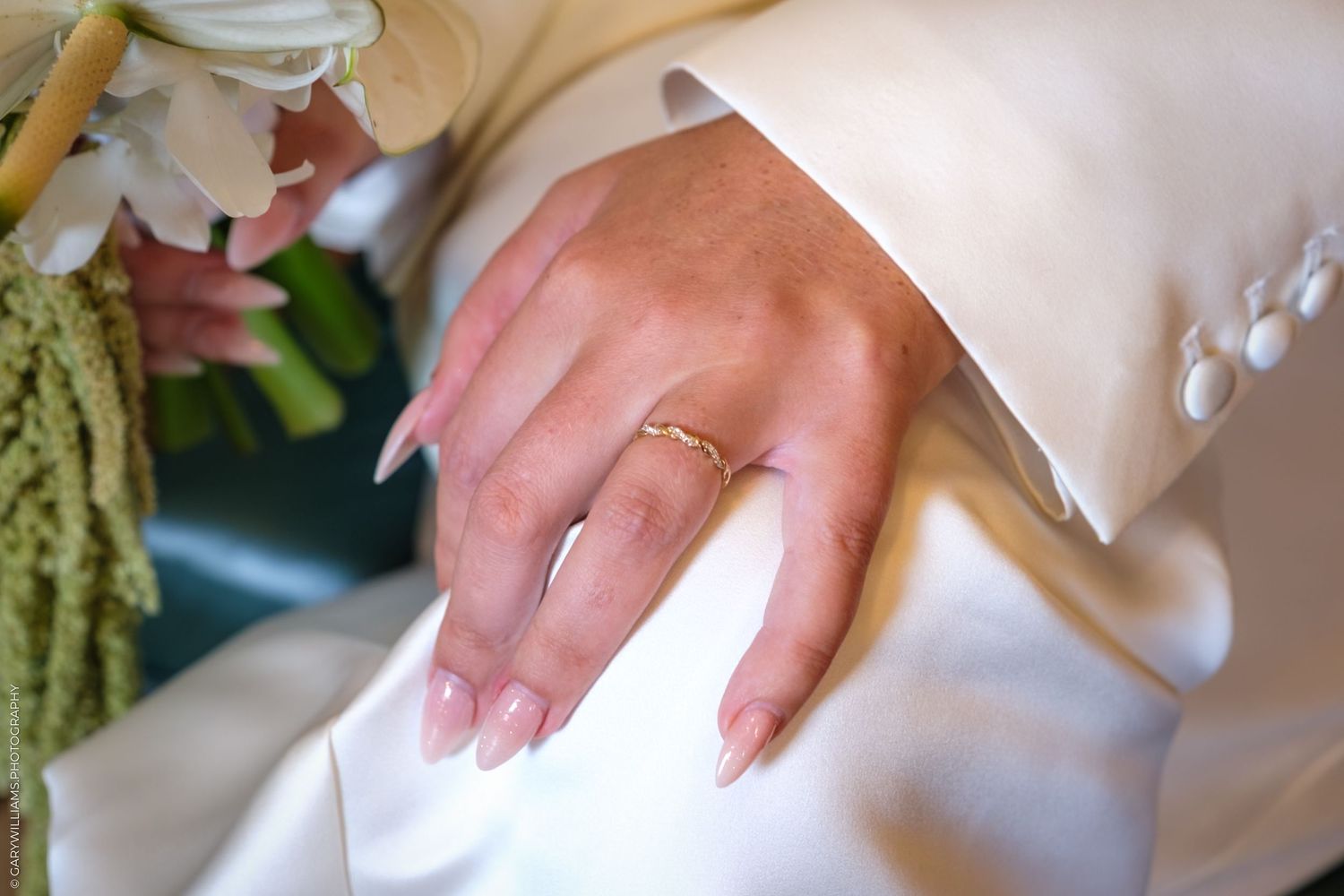 A delicate hand wearing a diamond engagement ring rests on white fabric, with soft floral accents in the background.