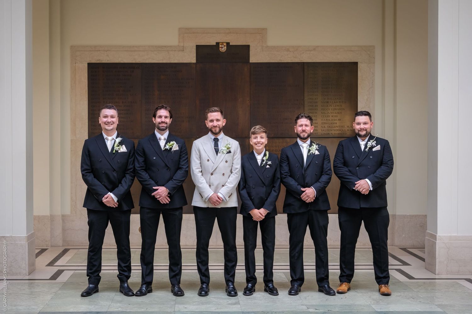 A group in matching suits stands in a line against a beige wall with columns.