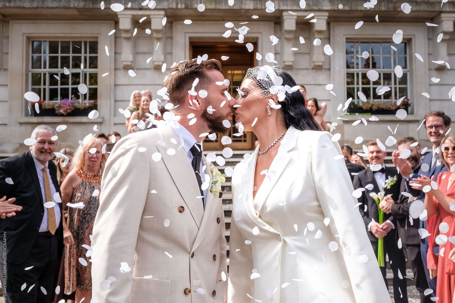 Wedding couple shares kiss under falling confetti petals while guests celebrate outside a historic building.