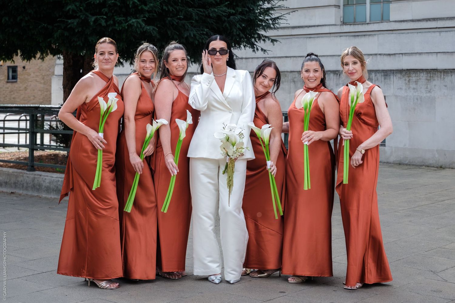 People in matching rust-colored dresses with green sashes stand in a row outside a building.