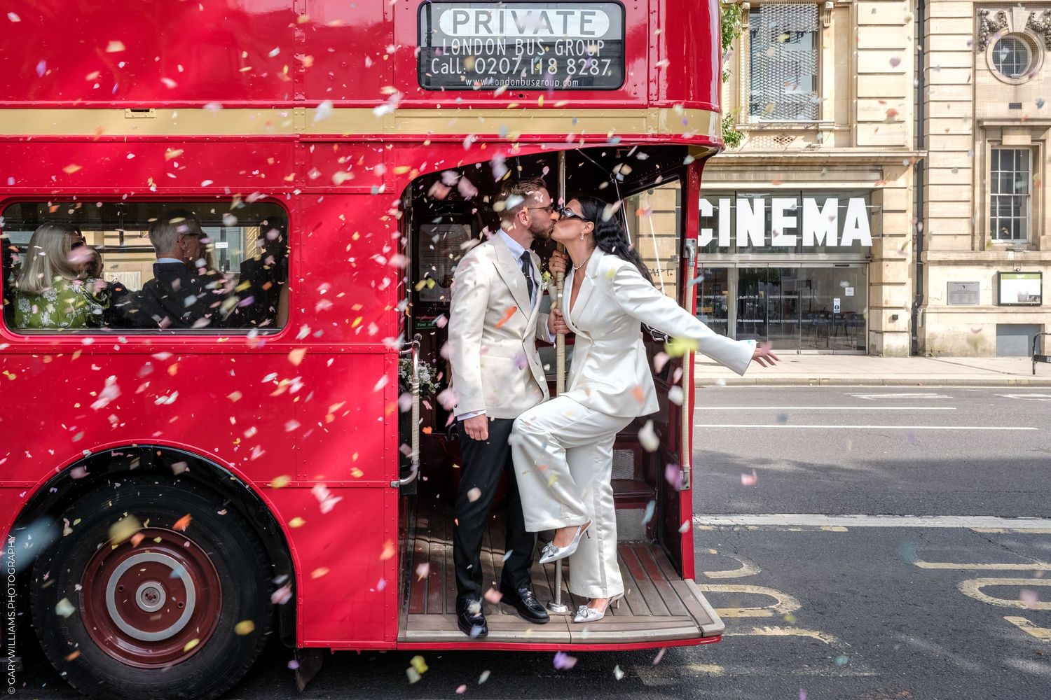 A wedding couple standing on the steps of a classic red London double-decker bus as confetti falls around them.