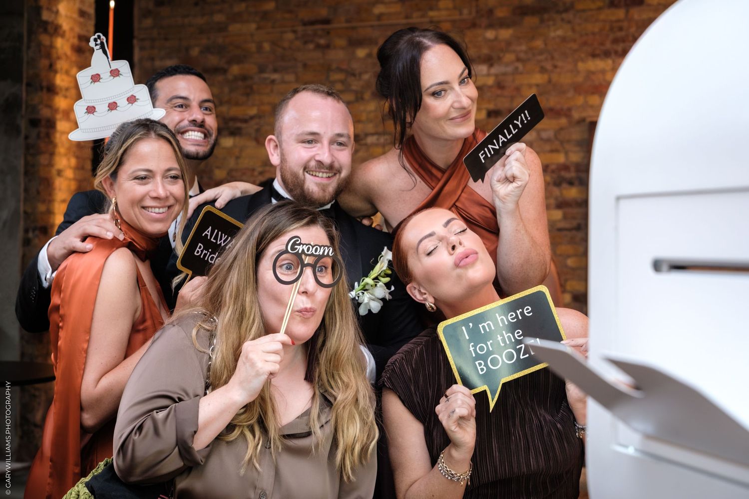 Small group poses casually with drinks and smiles at social gathering with exposed brick wall behind them.