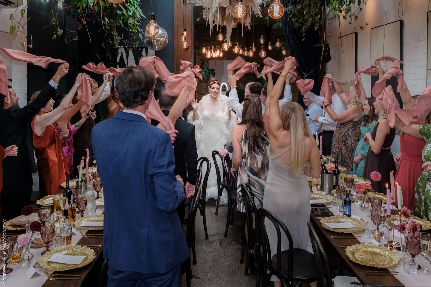 Wedding guests laughing and dancing at a festive indoor reception with pink decorations and table settings.