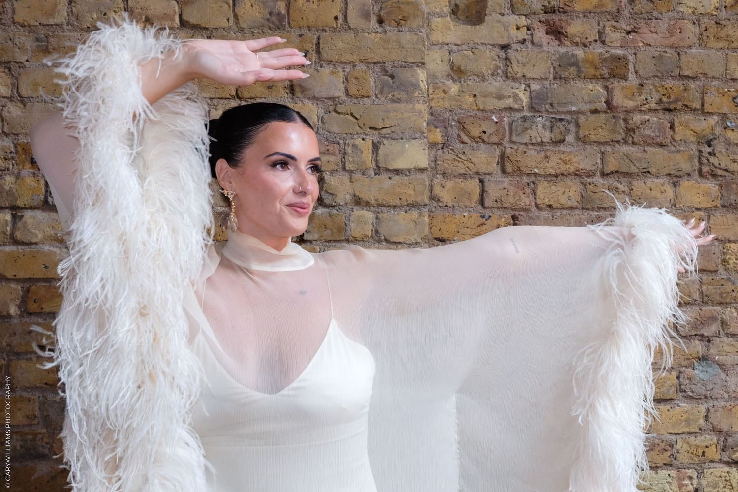 A fashion model in a white dress poses dramatically with feathered wings against a brick wall backdrop.