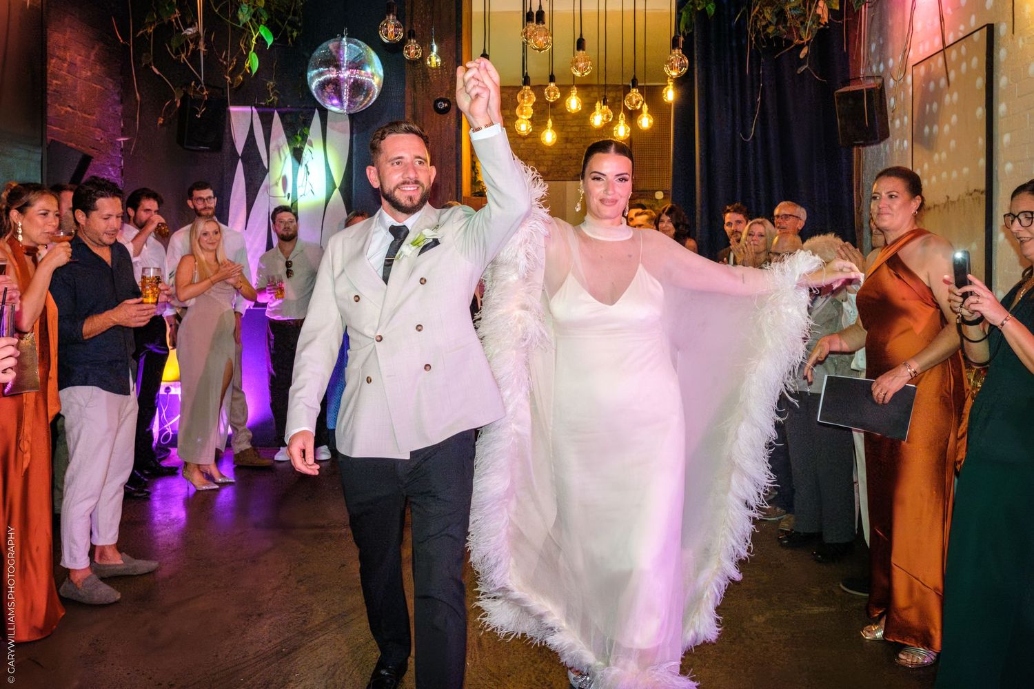 Joyful newlyweds make their grand entrance at a reception venue decorated with hanging lights and surrounded by guests.