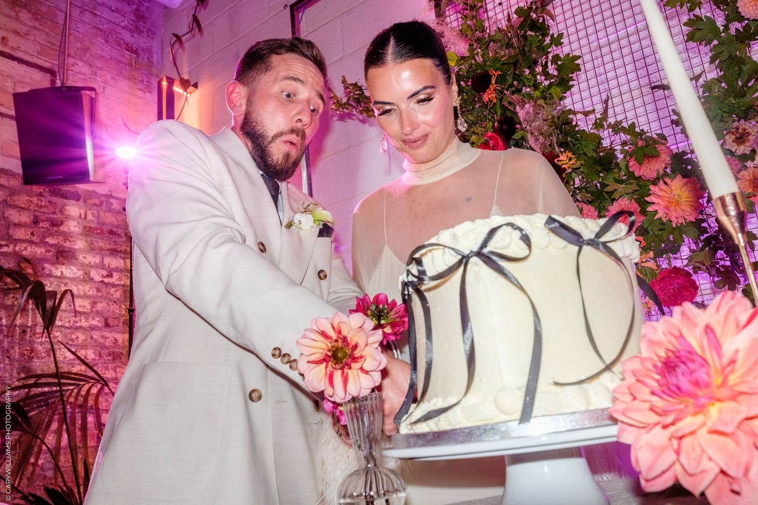 A couple cuts their white and black wedding cake together under pink lighting surrounded by floral decorations.