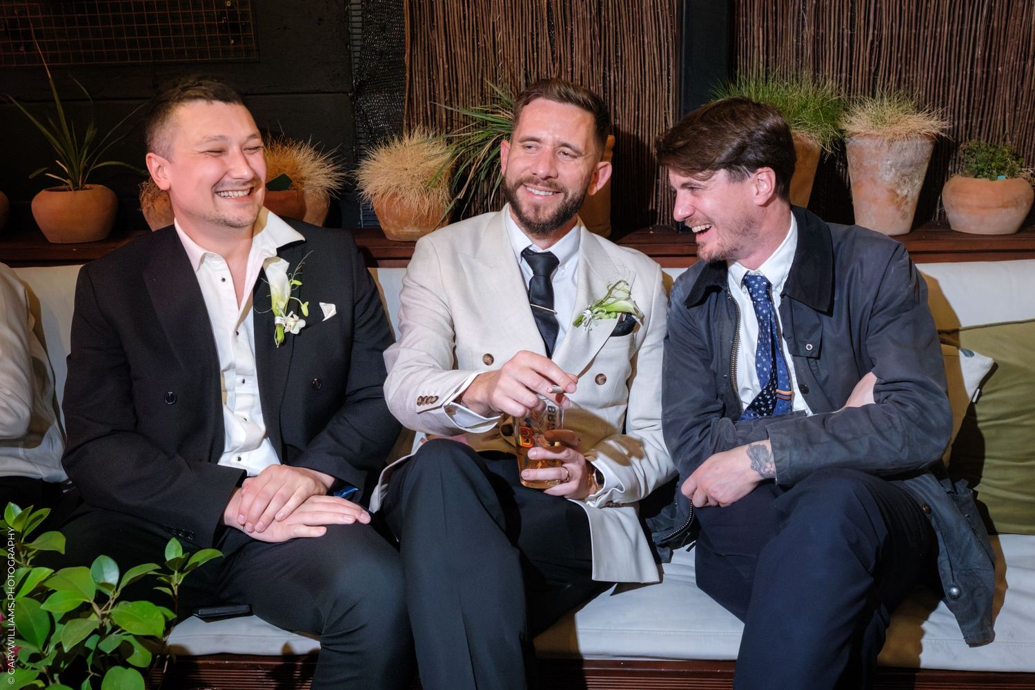 A group of laughing groomsmen in dark suits share a candid moment at a wedding celebration.