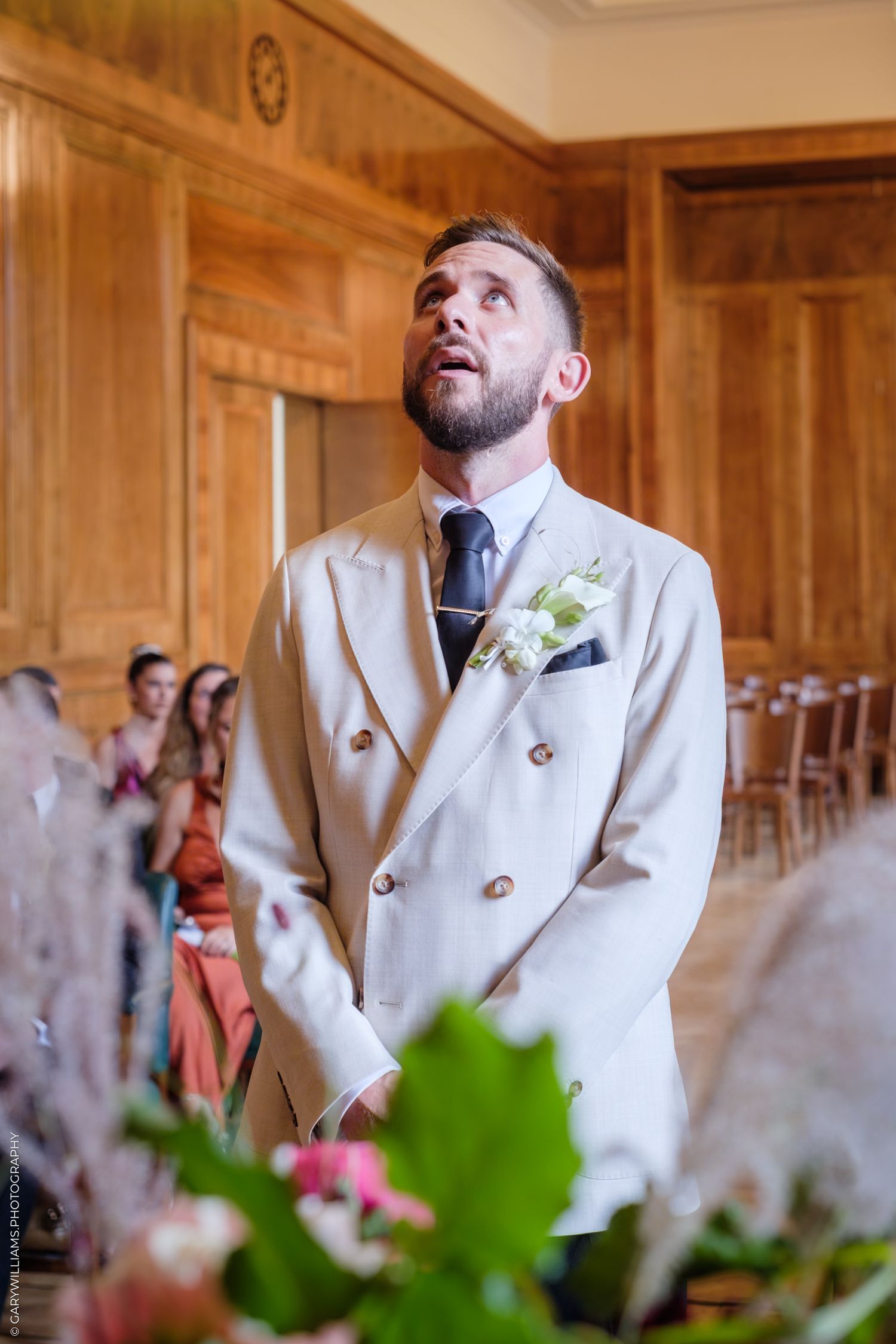 A person in a white suit jacket smiles with emotion during a special occasion in an elegant wood-paneled room.