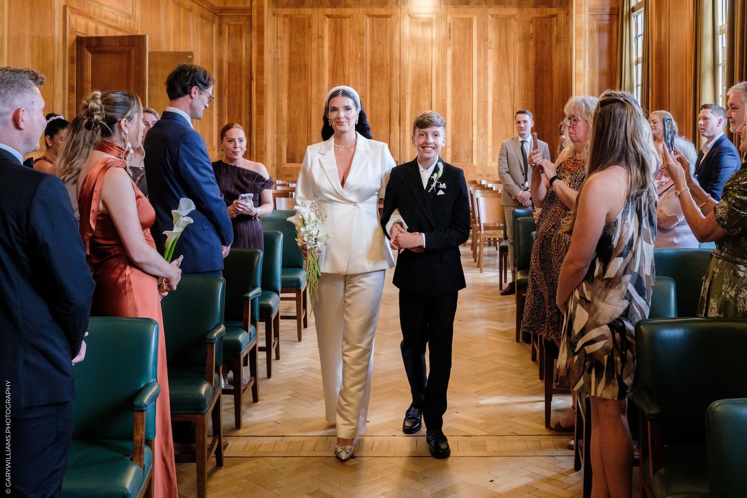 A processional at a formal indoor wedding ceremony with guests standing in rows of wooden chairs.