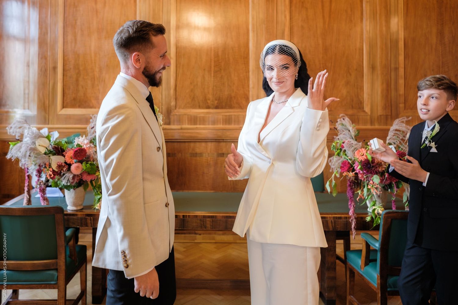 Wedding attendants in white and beige attire chat at a wooden-paneled indoor venue during a celebration event.