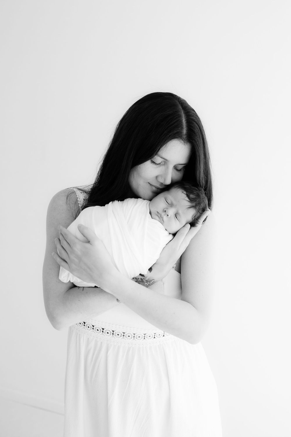 Mother in white dress tenderly embracing sleeping newborn baby in black and white portrait.