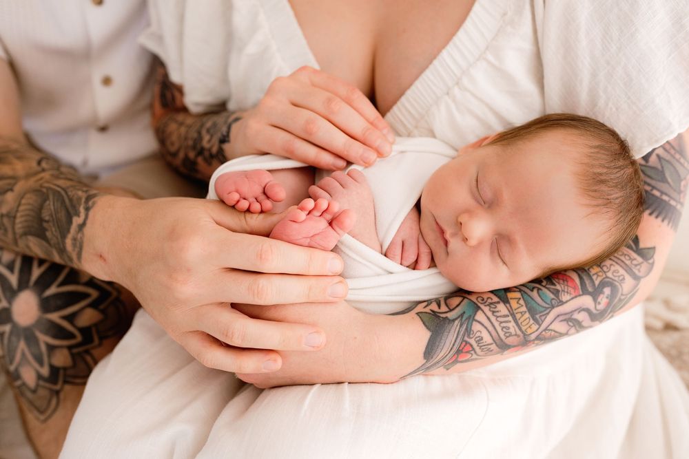 Tattooed hands gently holding sleeping baby's tiny feet against white fabric.