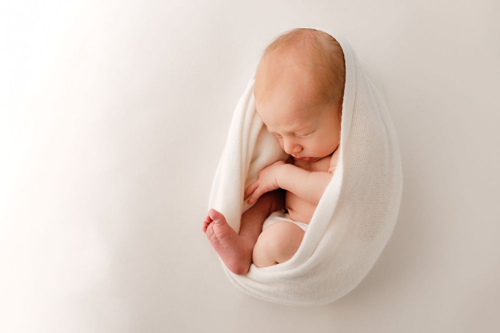 Artistic newborn photography with baby wrapped in white fabric against light background.
