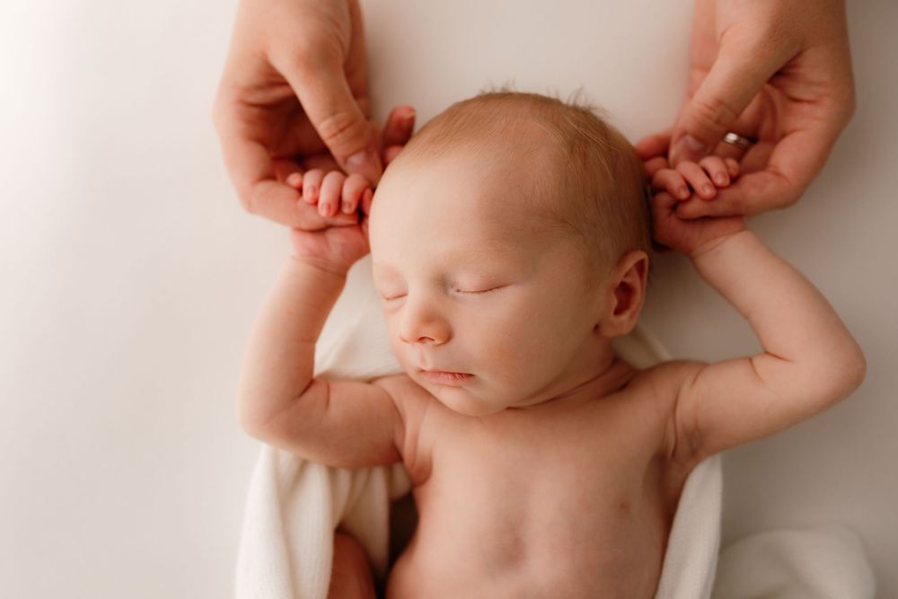 Sleeping newborn baby with arms raised above head while hands gently touch their ears on white background.