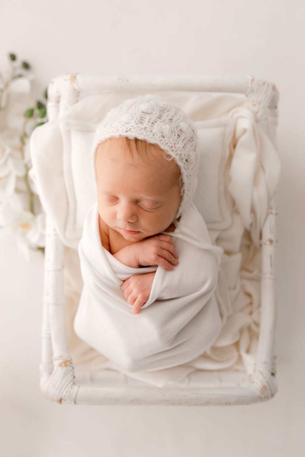Sleeping newborn in white bonnet rests on textured cream blanket during professional photo shoot.