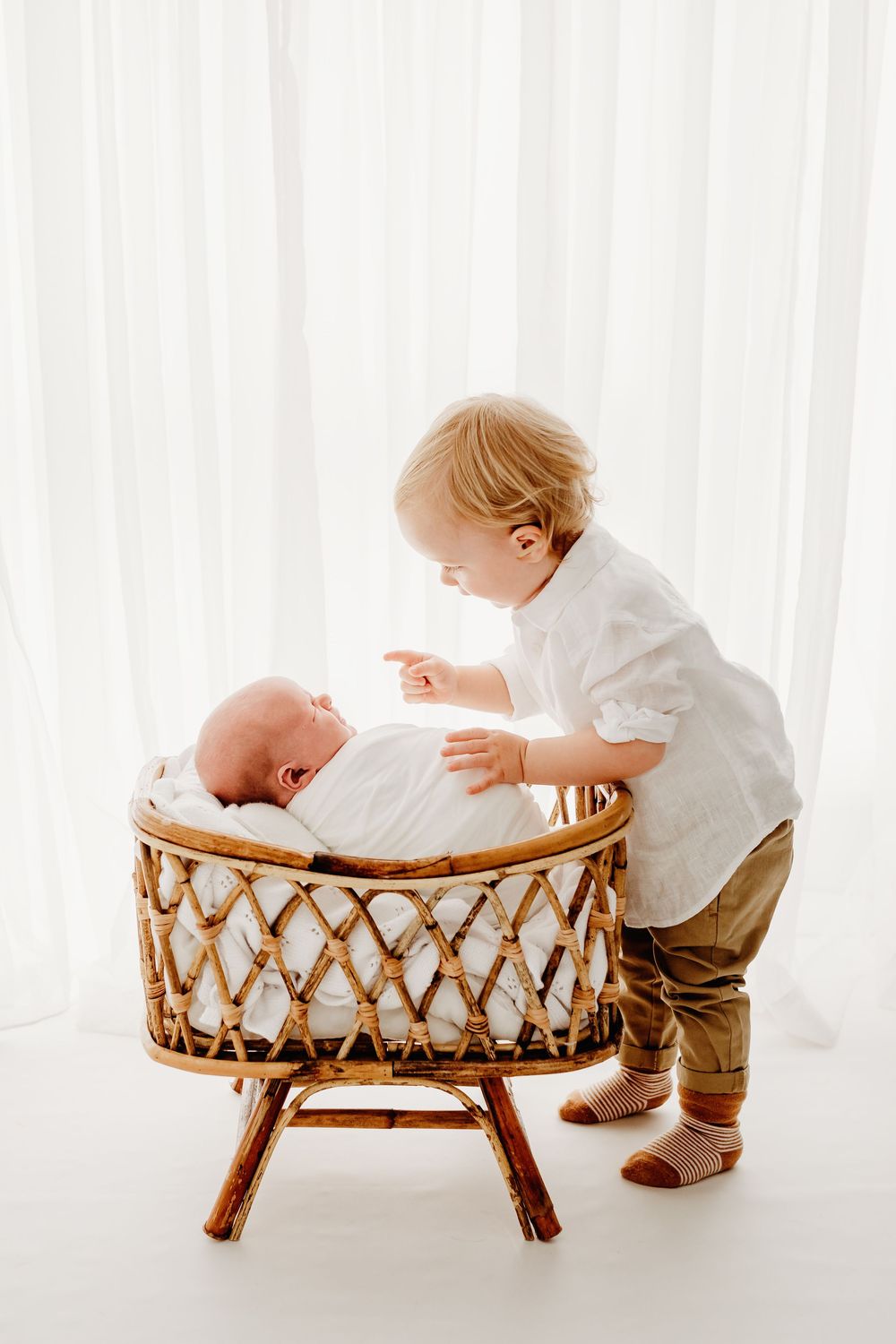 Toddler in white outfit peering into wicker bassinet during studio photo session.