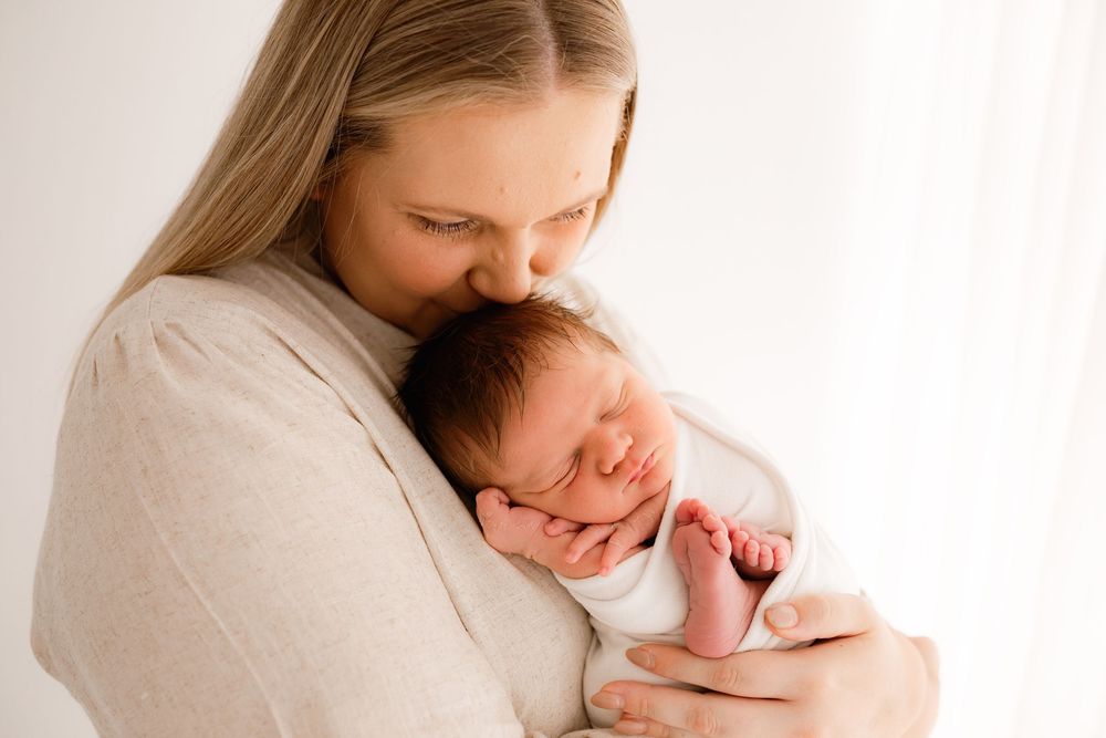 Mother in white dress holding and kissing sleeping newborn baby against bright window.