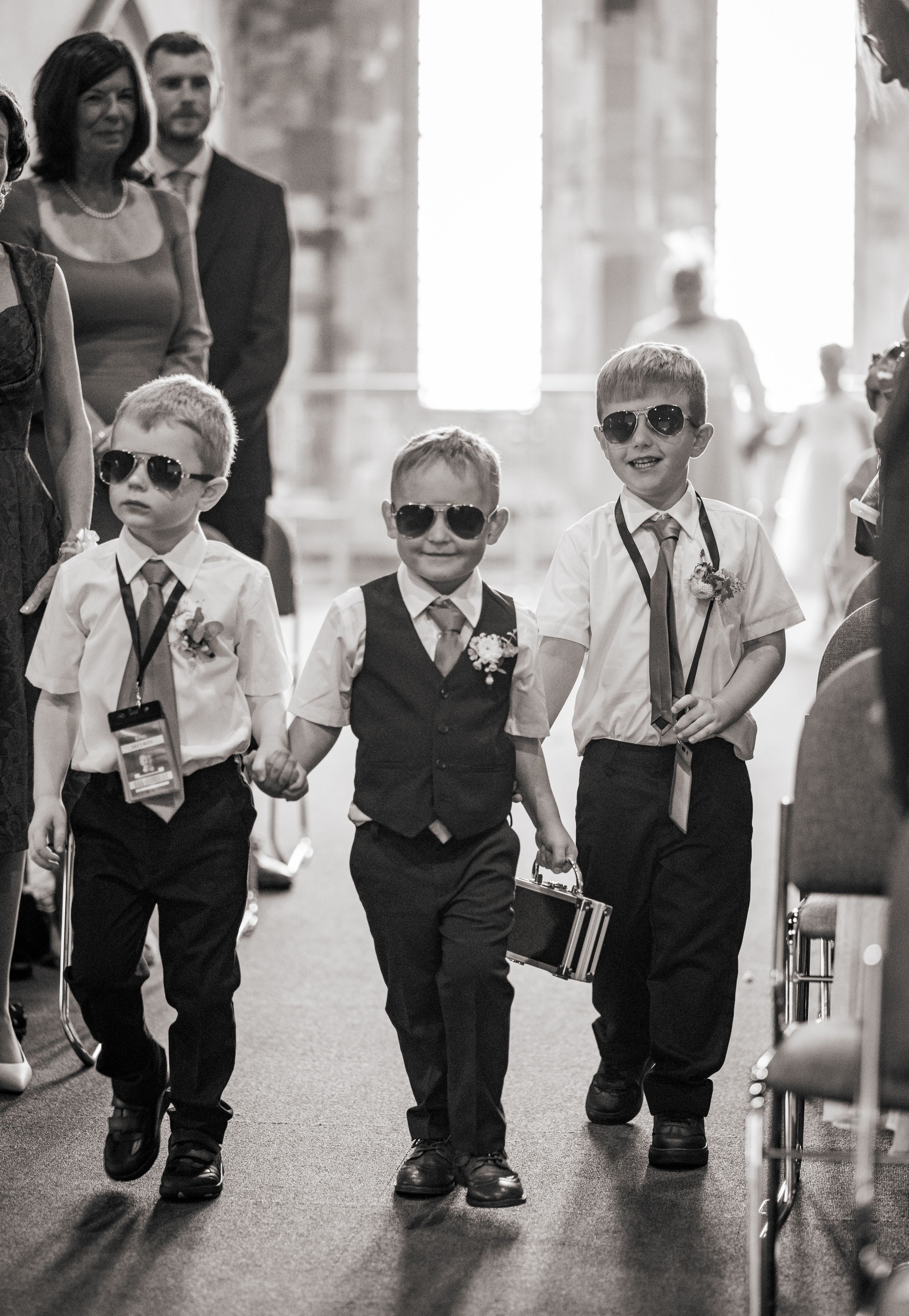 A group of young boys wearing sunglasses and formal attire walking in formation during a wedding ceremony.