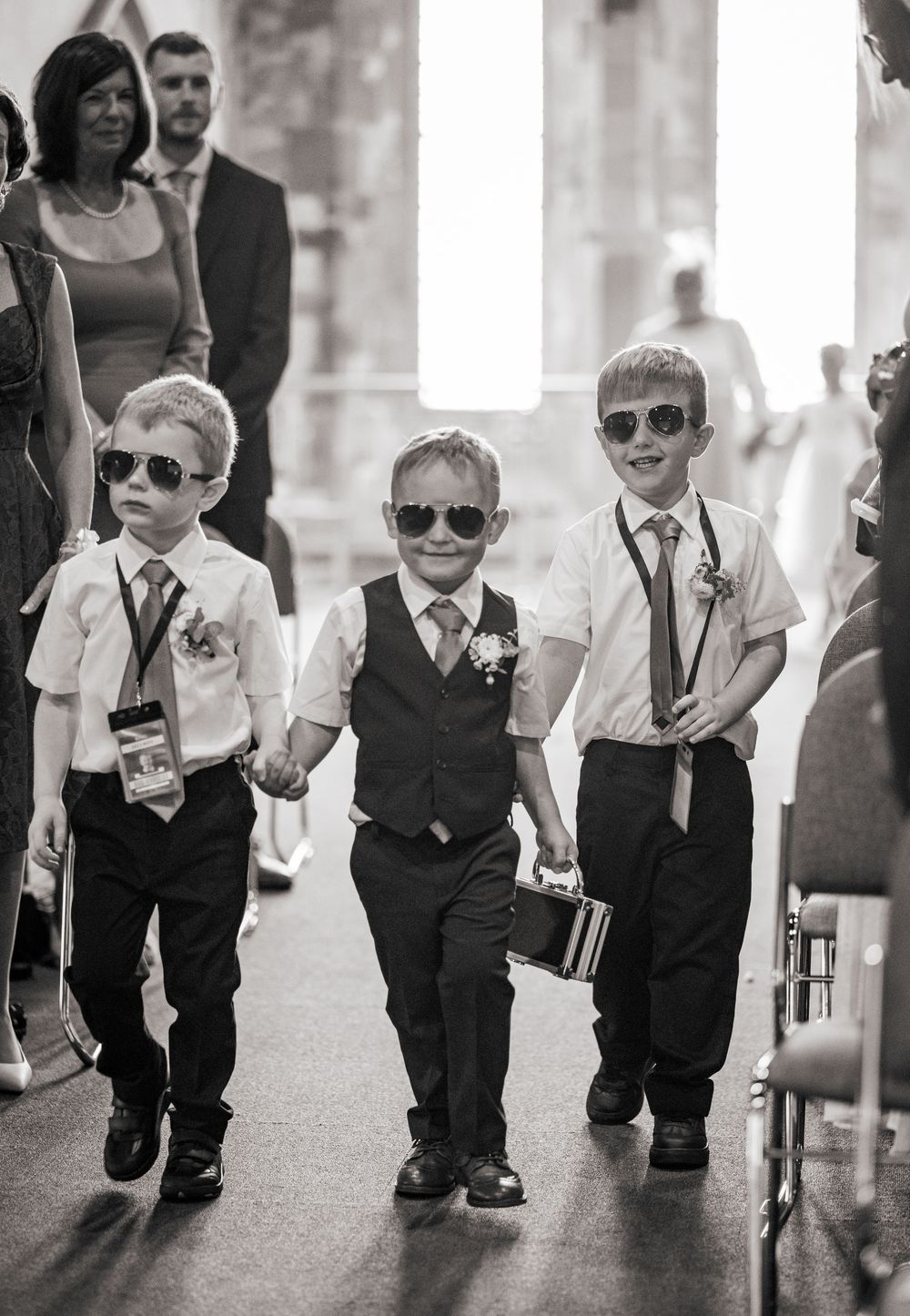 A group of young boys wearing sunglasses and formal attire walking in formation during a wedding ceremony.