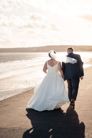 A bride in a flowing white gown and groom in dark suit walk along a serene beach at sunset.