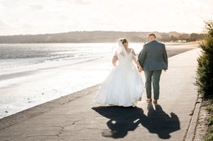 A couple in wedding attire walk along a scenic beach with their shadows cast on the sand at sunset.