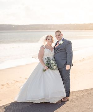 Couple in wedding attire embracing on a scenic beach during golden hour sunset.