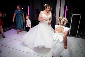 A bride in a white ballgown dances with a flower girl in white on an illuminated dance floor at a wedding reception.
