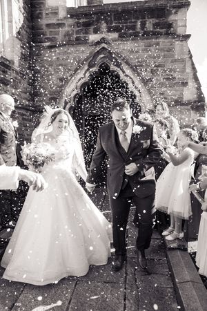 Confetti fills the air as newlyweds exit a gothic church building in black and white photography.