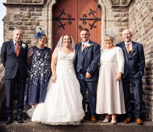 A wedding party stands in a line against a rustic stone wall with red doors in the background.