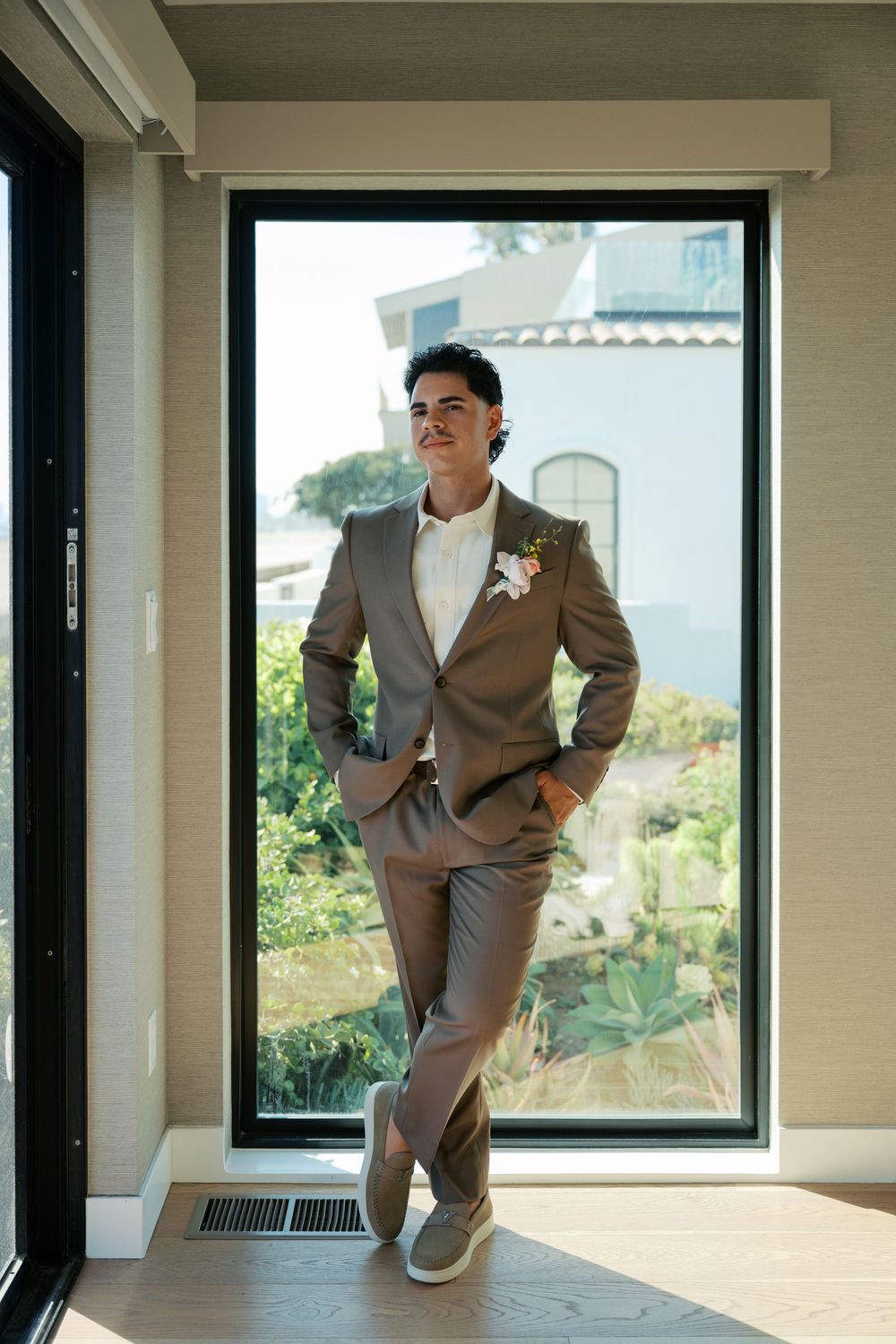 Person in a tan suit and white sneakers poses in front of a window in a series of playful fashion shots.