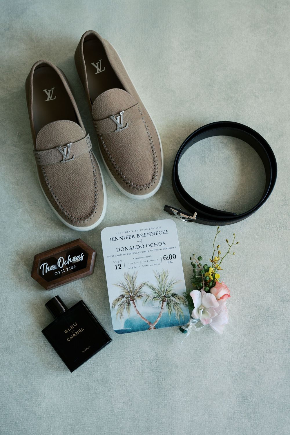Wedding details flatlay with Louis Vuitton loafers, sunglasses, invitations, and tropical floral accents on grey surface.