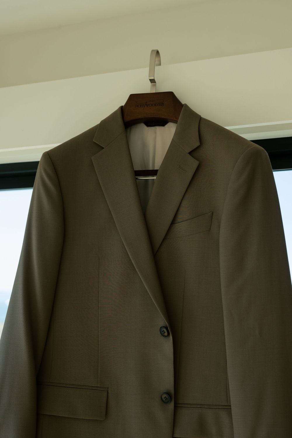 A gray business suit jacket hanging on display against a white wall in natural lighting.
