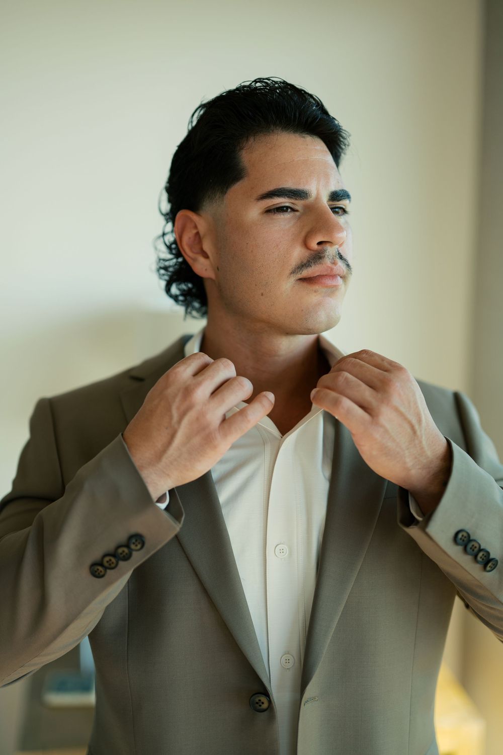 Person in a tan suit jacket and white shirt adjusts their collar while looking away in soft lighting.