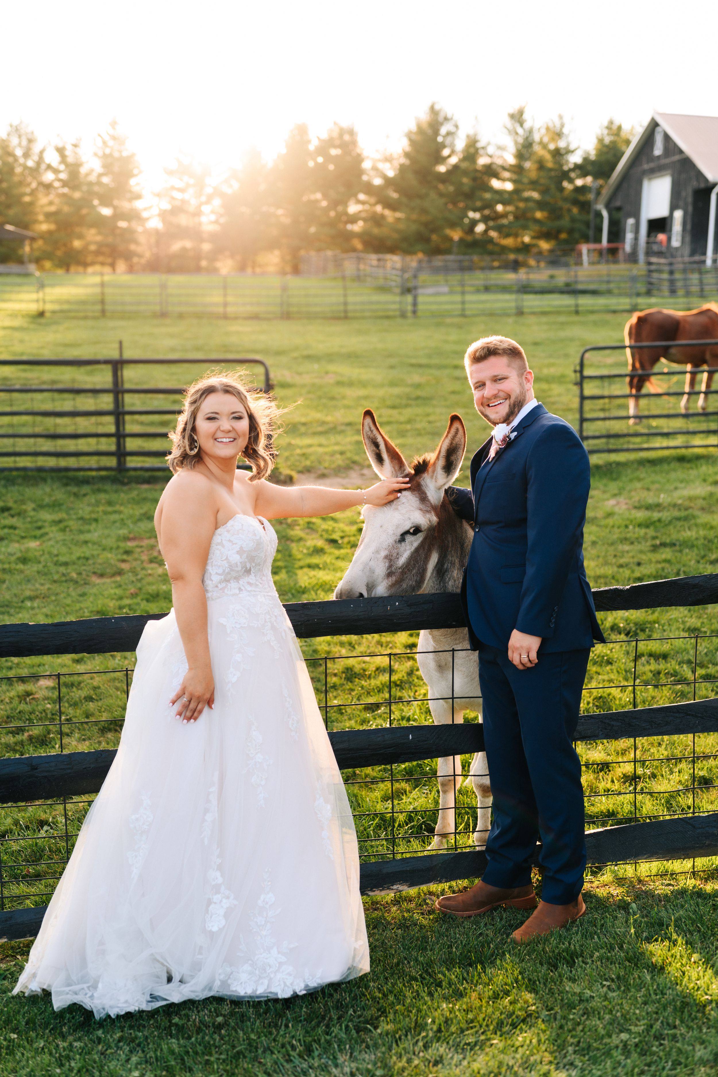 A couple takes playful wedding photos along a rustic wooden fence at sunset in a countryside farm setting.