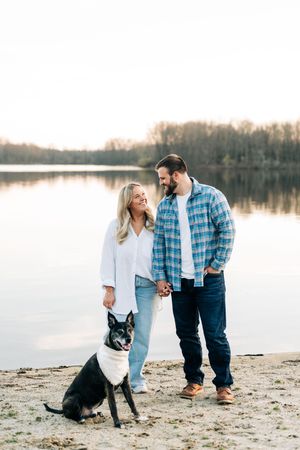 A couple shares a romantic moment with their black dog on a serene beach at sunset by a calm lake.