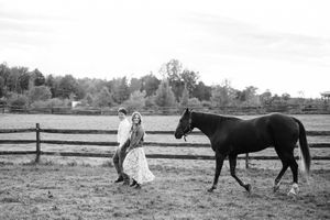 A graceful figure in a flowing dress stands in a field near a beautiful black horse by a wooden fence.