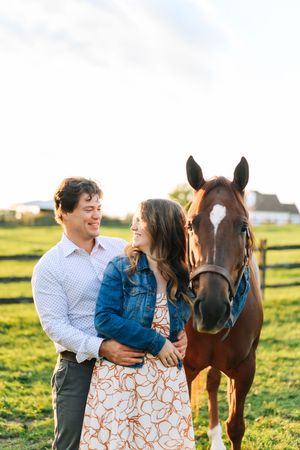 Couple shares romantic moments with their horse in a sunny pasture with wooden fencing at sunset.
