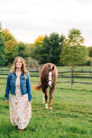 A brown horse grazes in a fenced pasture beside someone in a denim jacket and floral dress during autumn.