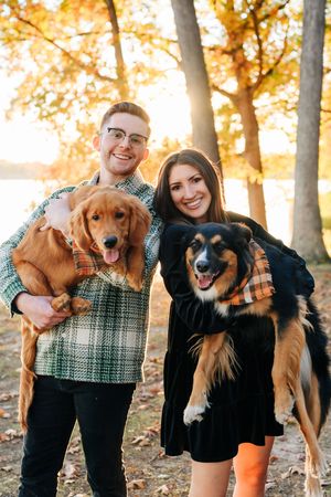Four smiling dogs being held up by their owners during a golden autumn sunset in the woods with fall foliage.