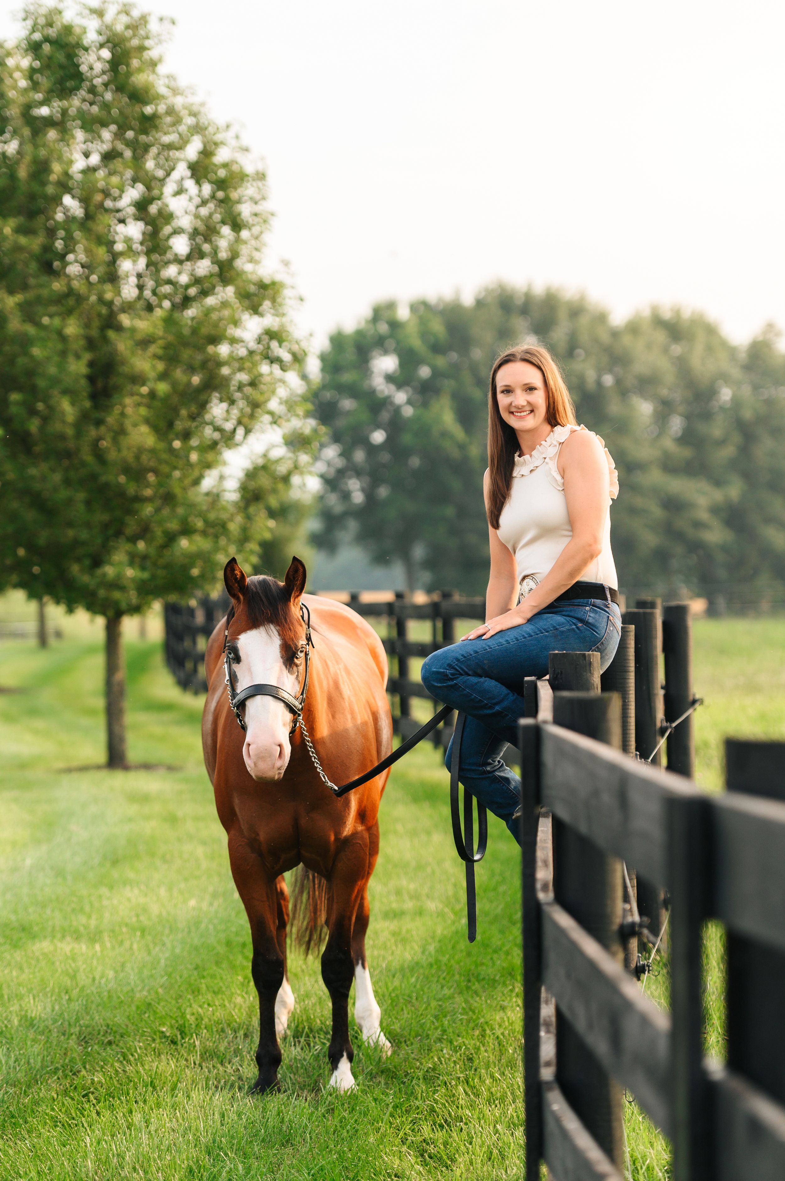 A woman sits on a black fence in blue jeans next to a brown and white paint horse in a green pasture.