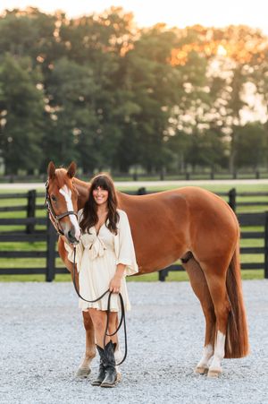 A chestnut brown horse stands with a rider in white clothing on a gravel arena at sunset.
