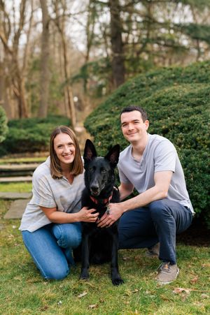 A loving couple poses outdoors with their beautiful black German Shepherd dog on green grass near manicured shrubs.