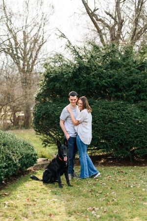 A couple shares a tender outdoor moment with their black dog among evergreen bushes in autumn.