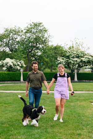 A couple walks their Border Collie dog on a lush green lawn with blooming trees in the background on a sunny day.