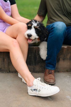 Two people sitting down with a small black and white dog between them wearing casual shoes and jeans.